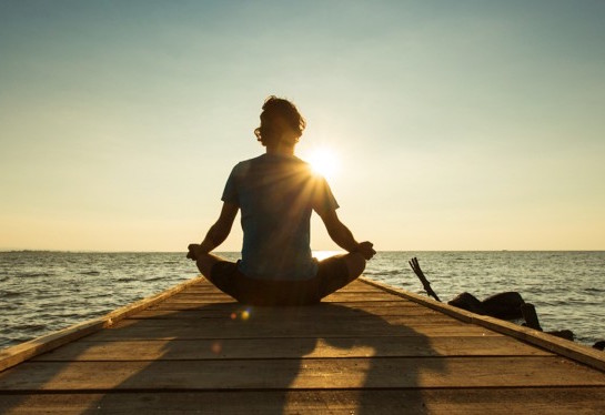 Young Man Sitting And Meditating On A Pontoon By A Lake At The Sunrise